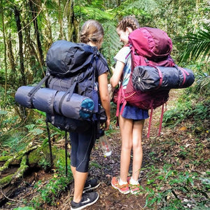 Two young females wearing backpacks with roll mats on hiking trail in rainforest