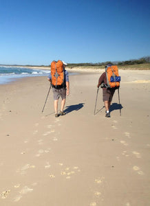Two males wearing full hiking packs walking on the beach
